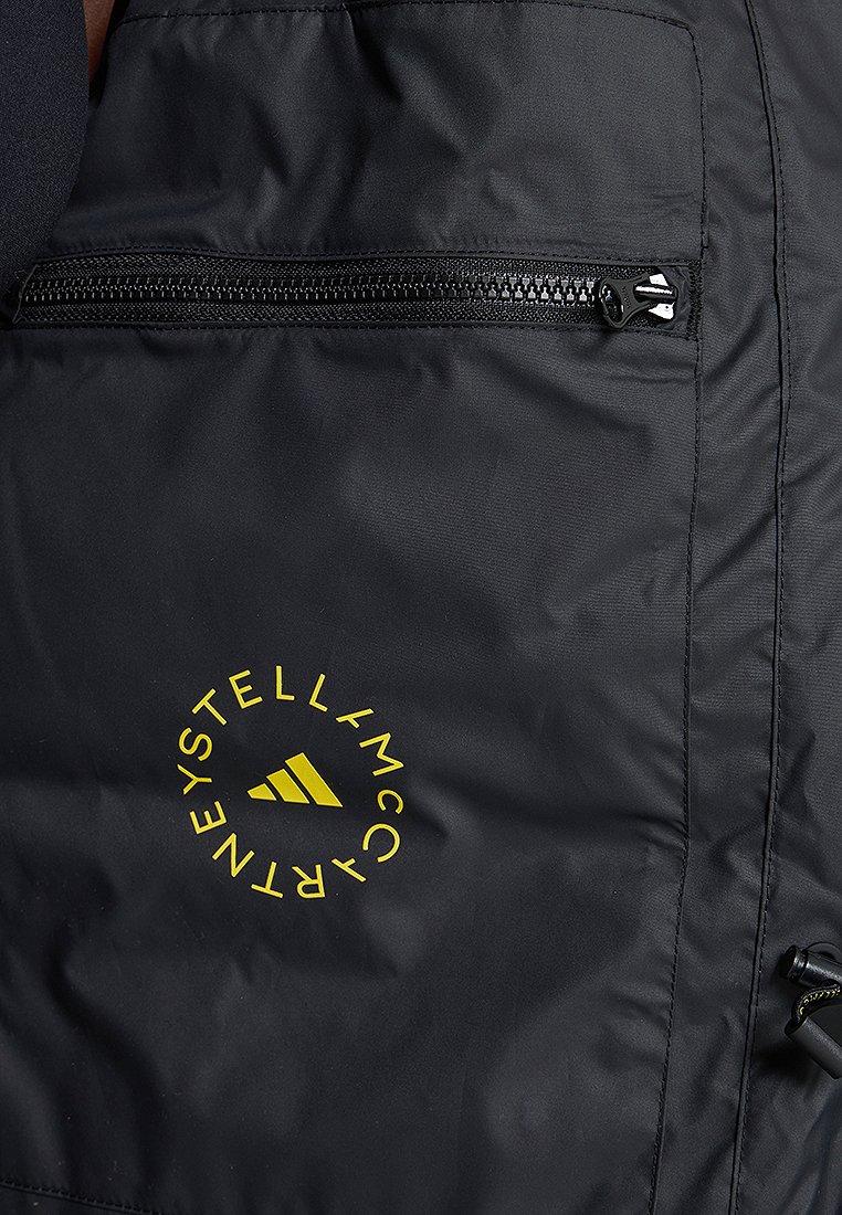 Schwarze Jacke mit einer Reißverschlusstasche, glatter Textur, auffälligem gelbem runden Logo mit Text und einem Design-Element mit drei Streifen.
