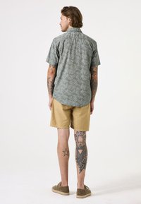 Kurzärmliges, hellgrünes Hemd mit Blattmuster, kombiniert mit sandfarbenen Shorts. Tattoo-Details an den Beinen sichtbar.