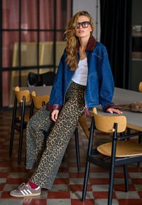 Džínová bunda s hnědým límcem, bílá košile a široké kalhoty s leopardím vzorem. Outfit doplňují hnědé a bílé tenisky.