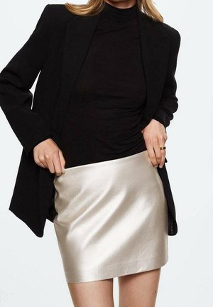 Femme portant un col roulé noir et un blazer ajuste une mini-jupe argentée brillante, visible des épaules jusqu'au milieu des cuisses sur un fond uni.