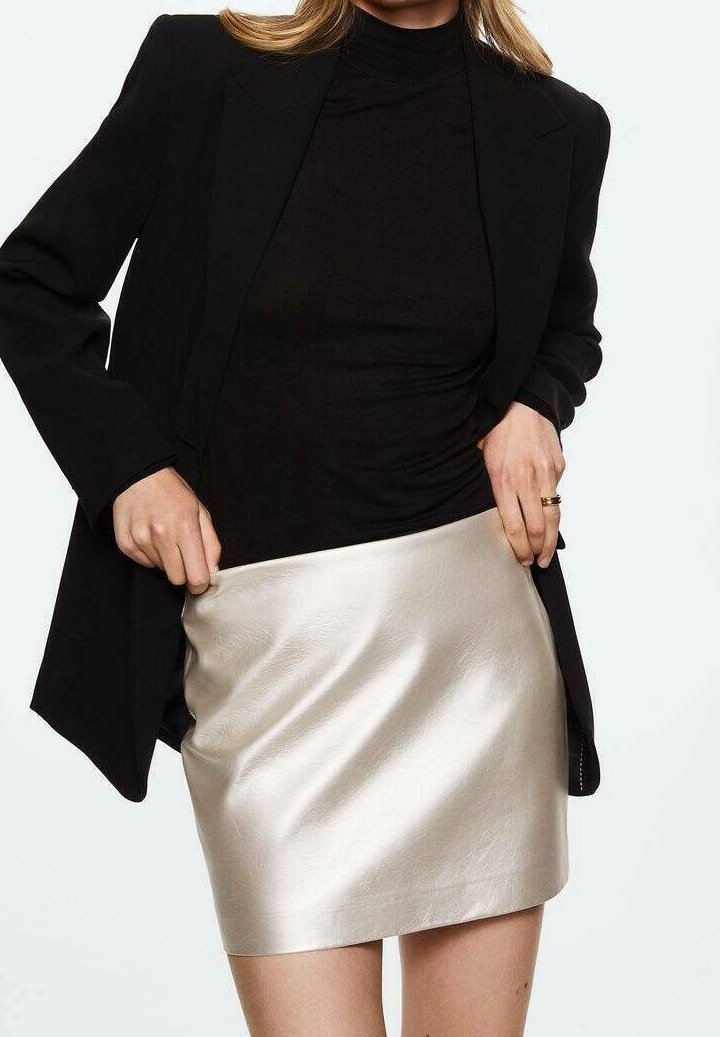 Femme portant un col roulé noir et un blazer ajuste une mini-jupe argentée brillante, visible des épaules jusqu'au milieu des cuisses sur un fond uni.