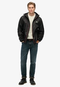 Joven con una chaqueta negra con capucha sobre un suéter blanco, jeans azules y botas negras, de pie con las manos en los bolsillos frente a un fondo blanco.