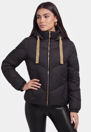 Frau trägt eine schwarze Steppjacke mit goldfarbenem Reißverschluss und Kapuzenkordeln, kombiniert mit schwarzen glänzenden Leggings, steht mit einer Hand in der Hüfte.