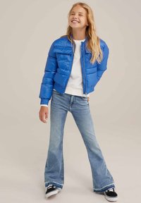 Kék puffertakos kabát, elöl cipzárral és bordás mandzsettákkal, világoskék, trapéz fazonú farmernadrággal és fekete sportcipőkkel párosítva. Puha tapintású, könnyű kialakítás.