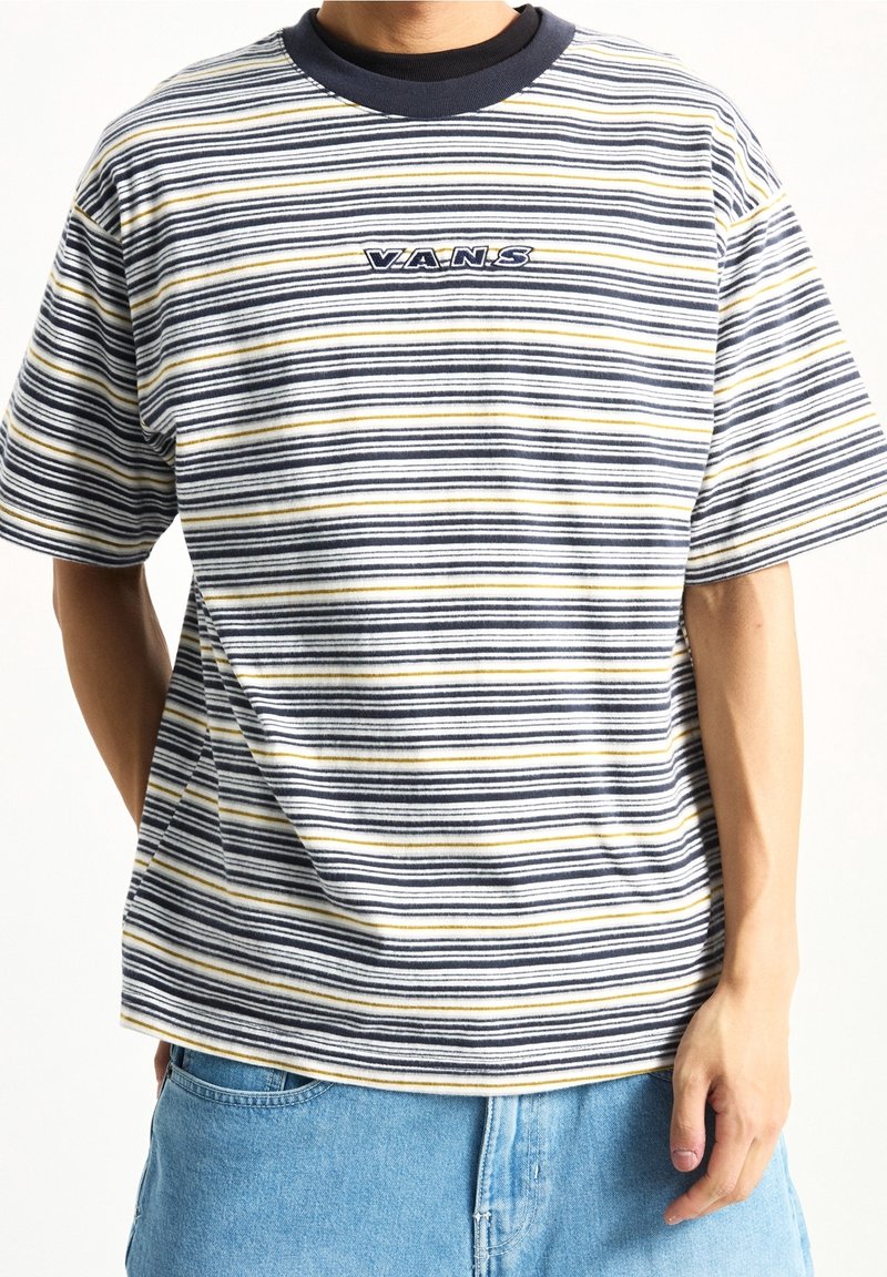 Personne portant un t-shirt Vans rayé avec des lignes noires, blanches et jaunes, associé à un jean denim bleu clair.