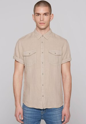 Junger Mann mit kurz geschorenem Haar, trägt ein beiges kurzärmeliges Hemd mit Brusttaschen und blaue Jeans, steht vor einem schlichten Hintergrund.