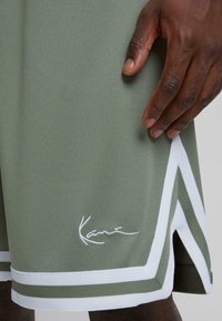 Olive green mesh basketball shorts with white striped accents and a stitched logo at the hem. Features side slits for ease of movement.