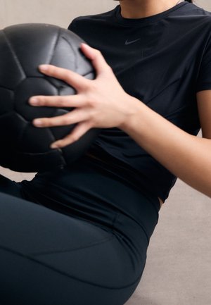 Personne vêtue de vêtements de sport noirs tenant un ballon médicinal noir, assise sur un sol clair.