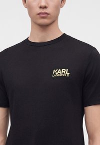 Homme portant un t-shirt noir avec un petit logo jaune "KARL LAGERFELD" sur la partie gauche de la poitrine, fond clair uni.