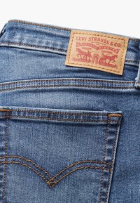 Jeans en denim avec une finition bleue, arborant une étiquette de marque en cuir marron à la taille et des coutures orange contrastantes.