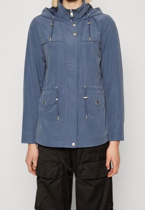 Persona che indossa una giacca blu con cappuccio, tasche frontali e coulisse, abbinata a pantaloni neri con ampie tasche cargo.