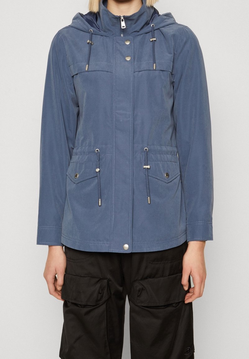 Personne portant une veste bleue à capuche avec poches avant et cordons, associée à un pantalon noir avec grandes poches cargo.