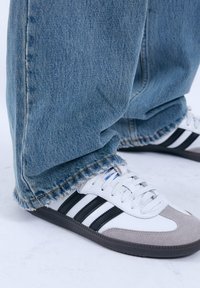 Witte sneakers met zwarte strepen, met een grijze suède teen. Gecombineerd met lichtblauwe denim jeans, gescheurd aan de zoom, wat textuur toont.