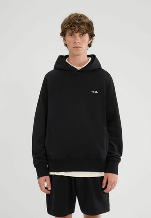 Jonge man met krullend haar draagt een zwarte ellesse-hoodie en zwarte korte broek, staand tegen een effen lichte achtergrond.