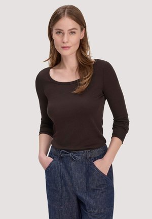 Junge Frau mit langem hellbraunem Haar, die ein schwarzes Shirt mit rundem Ausschnitt und hoch taillierte blaue Jeans mit den Händen in den Taschen trägt.