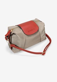 Bolso de nailon beige con una solapa y correa de cuero rojo. Presenta un cierre dorado y detalles de marca. Forma compacta y redondeada con textura suave.