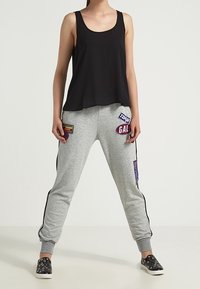 Canotta nera senza maniche abbinata a pantaloni da tuta grigi con toppe colorate, strisce laterali e scarpe slip-on nere con motivo floreale.