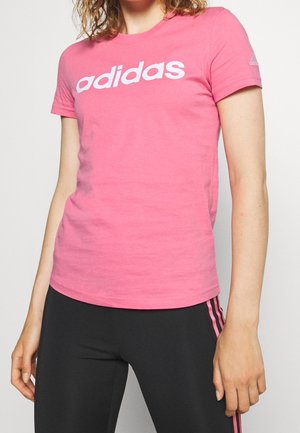 Kvinde iført en pink Adidas t-shirt og sorte leggings med pink striber, står mod en ensfarvet lys baggrund.