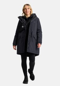 Donkerblauwe parka met een rits aan de voorkant, capuchon en meerdere zakken. Gepaard met zwarte leggings en sneakers. Compacte zwarte tas schuin over de schouder gedragen.