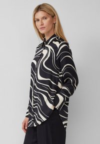 Donna con lunghi capelli biondi, che indossa una camicia a bottoni con motivo astratto in bianco e nero e pantaloni neri, in piedi contro uno sfondo grigio.