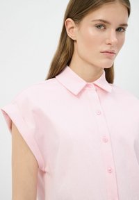 Camicia col collare di colore rosa chiaro con maniche corte arrotolate, con chiusura a bottoni e una sottile texture. Presenta sei bottoni e una vestibilità rilassata.