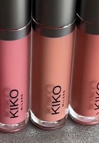 Tre KIKO Milano lipglosserør i rosa, fersken og korallfarger med sølvhetter på en teksturert grå overflate.