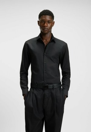Black button-up shirt with a subtle textured pattern, long sleeves, and a pointed collar, paired with loose-fitting black trousers and a belt.