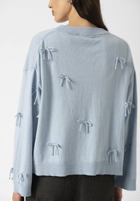 Light blue sweater with decorative bows on the back, worn by a woman with long hair. Focus on textured fabric and unique design details.