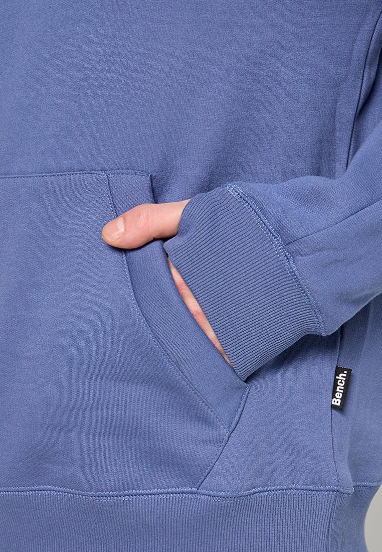 Sudadera azul con un bolsillo frontal, puños y dobladillo acanalados. Presenta una pequeña etiqueta de logo negra en el lado, hecha de tela suave.