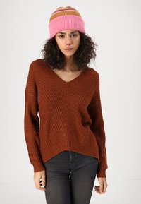 Bruine gebreide trui met een V-hals en een losse pasvorm, gecombineerd met een meerkleurige gestreepte gebreide muts. De textuur is chunky en comfortabel.