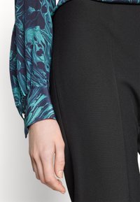 Blus med långa ärmar i blå och grön marmorering, med knäppta manschetter, parat med svarta kroppsnära byxor, uppvisad på en hand och arm.