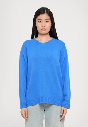 Blå strikkegenser med rund hals, lange ermer og ribbestrikkede mansjetter. Bærers over lyse jeans, med en avslappet passform og glatt tekstur.