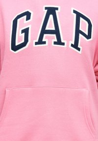 Ροζ φούτερ με γράμματα "GAP" σε μπλε ναυτικό και λευκό χρώμα, περιγραμμένα, πάνω από μια εμπρόσθια τσέπη τύπου ποδιά.