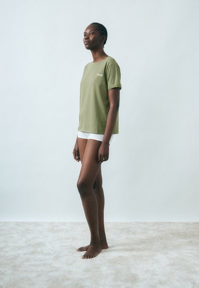 Femme debout pieds nus sur un tapis clair, portant un T-shirt vert olive et un short blanc, devant un fond uni gris pâle.