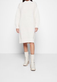 Robe-pull crème en maille épaisse avec manches longues et poignets côtelés, associée à des bottines beige brillant avec détail de sangle latérale.