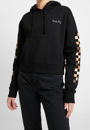 Persoon draagt een zwarte capuchonsweatshirt met een voorzak en een meerkleurig geruit patroon op de mouwen.