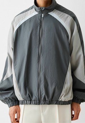 Homme portant une veste coupe-vent zippée grise et beige avec poignets et ourlet élastiques, debout devant un fond uni.