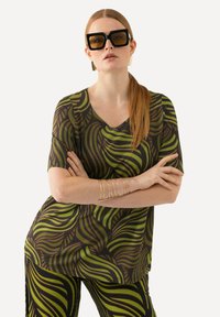 Top a V con maniche corte in tessuto scuro, caratterizzato da motivi di foglie verdi e marroni a spirale. Il modello indossa occhiali da sole quadrati grandi e bracciali dorati.