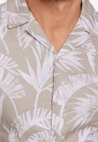 Chemise à manches courtes en tissu beige avec imprimé de feuilles de palmier blanches, dotée d'un col camp et d'une patte de boutonnage. Design texturé et léger.