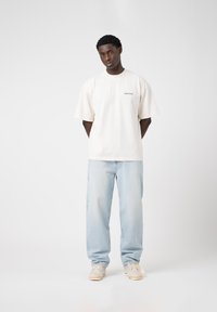 Light beige oversized t-shirt with "EIGHTYFIVE" embroidered on the chest, paired with light blue straight-leg jeans and beige sneakers.