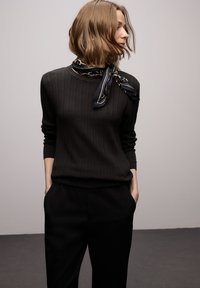 Maglione nero a coste con maniche lunghe, indossato con una sciarpa nera e oro con motivo. Abbinato a pantaloni neri che mostrano una vestibilità comoda.