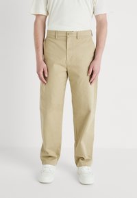 Beige chino's gemaakt van katoen; beschikt over een knoopsluiting en zijzakken; recht model; gedragen met witte sneakers.