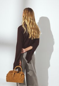 Brauner Langarmoberteil mit weiten Ärmeln, kombiniert mit hellgrauen Hosen. Eine handgefertigte Tasche aus Wildleder in Beige mit runden Griffen wird in einer Hand gehalten.