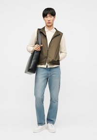 Νέος άνδρας με μπεζ μακρυμάνικο πουκάμισο, ανοιχτό πράσινο γιλέκο με φερμουάρ, ανοιχτό μπλε τζιν, λευκά αθλητικά παπούτσια, κρατώντας μια μεγάλη μαύρη τσάντα ώμου.