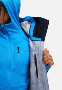 Die blaue Zip-Jacke verfügt über einen strukturierten Stoff, eine Kapuze und ein kontrastierendes graues Innenfutter. Sie hat Reißverschlusstaschen und eine Markenkennzeichnung auf der Vorderseite.