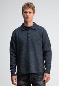 Dunkelgrauer Wollpullover mit Kragen, vier Knöpfen und langen Ärmeln. Strukturierter Stoff mit entspannter Passform, über einem schwarzen T-Shirt und Jeans getragen.