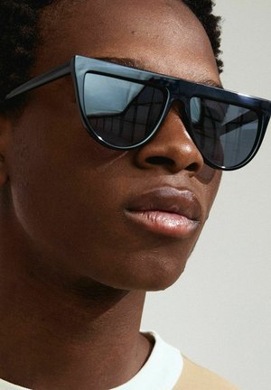 Black oversized sunglasses with angular frames, dark lenses, and glossy finish. Worn by a person with short hair, neutral shirt visible.