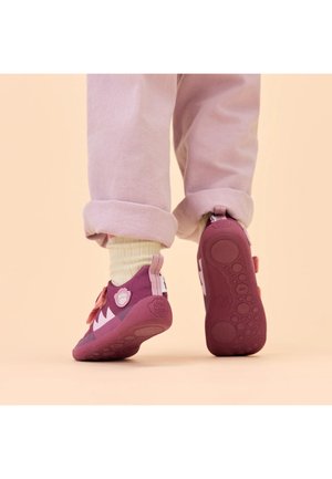 Kind draagt roze sneakers met witte sokken en opgerolde broek, loopt op een perzikkleurige ondergrond.