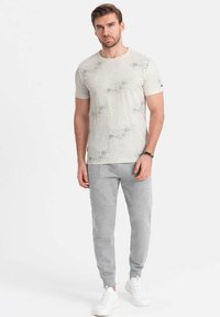 Camiseta de manga corta en color beige claro con motas oscuras dispersas, combinada con pantalones de chándal grises ajustados y zapatillas blancas. Diseño y ajuste sencillos.