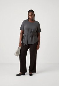 Graues kurzärmeliges Oberteil mit einer Schleifendetail am Bund, kombiniert mit schwarzen, weit geschnittenen Hosen und schwarzen Loafers. Hält eine kleine gemusterte Tasche.